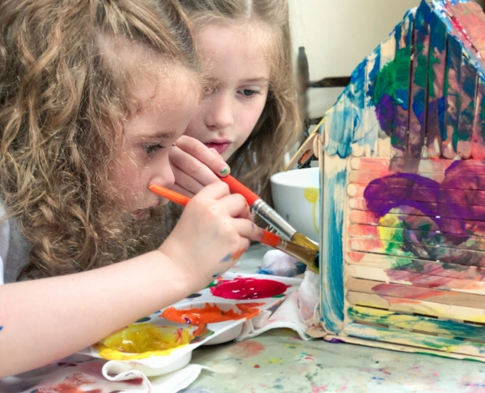 Two young children with curly hair focus intently while painting a wooden house model with bright, colorful paints.